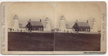 Load image into Gallery viewer, Antique card with two nearly identical images showing the original CHatham Lighthouses aka twin lights with the dark colored keepers building between them and a white fence upon which figures sit, lean, and stand with a grouping of seated figures in the grass in front of the fence. Photo was taken at a distance and the figures features are not discernable. 
